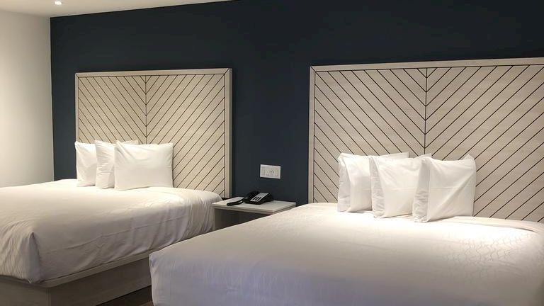 A modern hotel room featuring two neatly made beds with chevron-patterned headboards, and a sleek nightstand in between, against a dark blue accent wall.