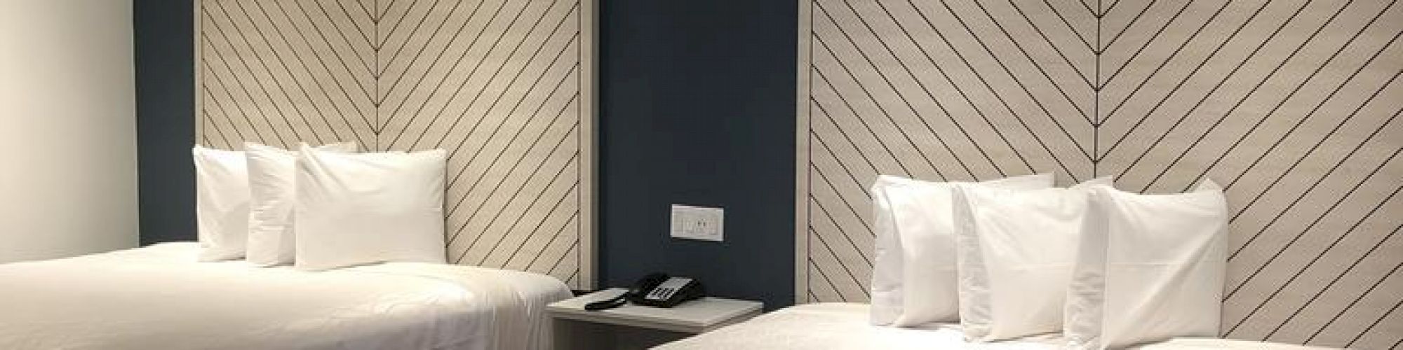 A modern hotel room featuring two neatly made beds with chevron-patterned headboards, and a sleek nightstand in between, against a dark blue accent wall.