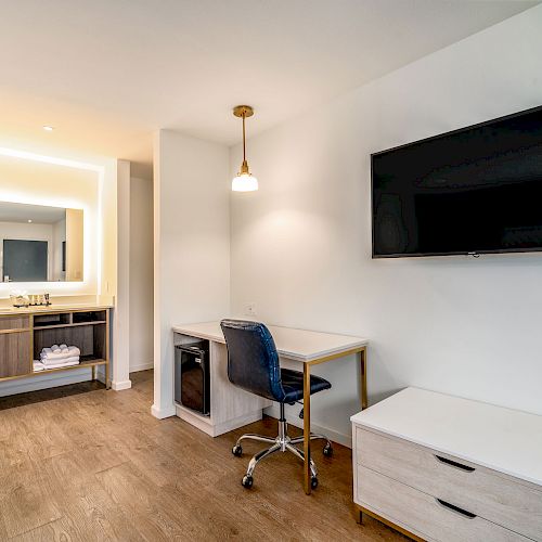 This image shows a modern hotel room with a desk, chair, wall-mounted TV, and a bathroom vanity area with illuminated mirror and storage.