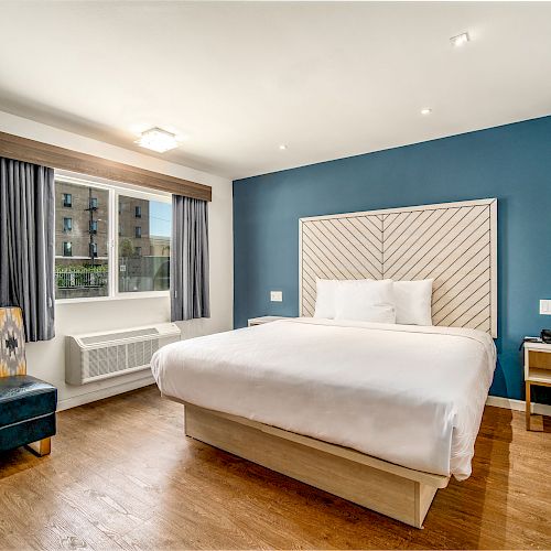 A modern hotel room with a large bed, bedside tables, a chair, and a window with curtains, featuring a blue accent wall.