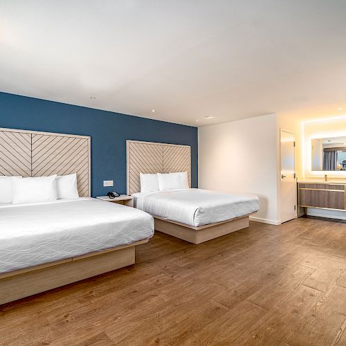 The image shows a modern hotel room with two neatly made double beds, a blue accent wall, wooden flooring, and a spacious bathroom area.