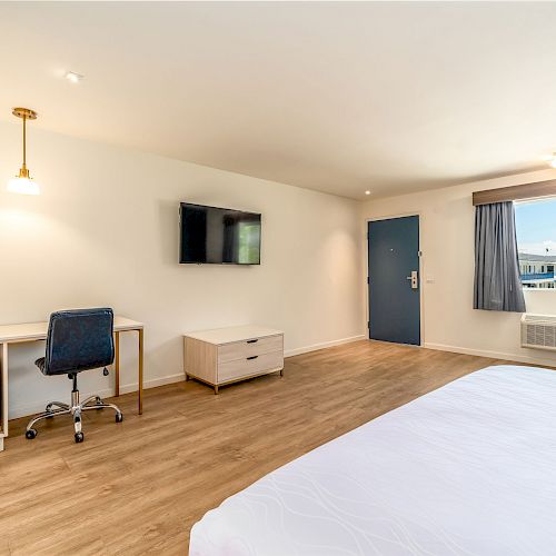 A modern hotel room with a bed, desk and chair, TV, mini fridge, air conditioner, window with closed curtains, and a blue door.