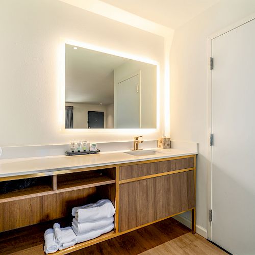 A modern bathroom with a lighted mirror, white sink, wooden cabinet, and neatly folded towels next to a closed door.