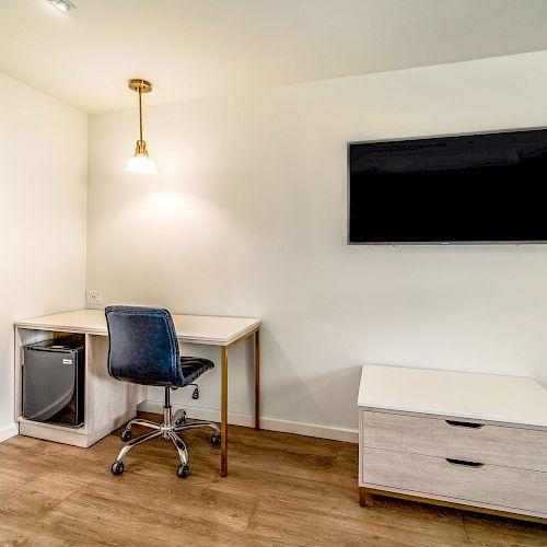 A minimalist room with a desk, chair, mini-fridge, wall-mounted TV, and a small cabinet, featuring wooden floors and a pendant light above the desk.