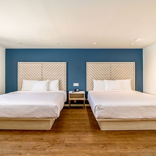 This image shows a clean, modern hotel room with two large beds, white bedding, a blue accent wall, and wooden flooring.