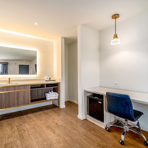 This is a modern interior with a bathroom vanity mirror and sink to the left, and a work desk with a blue chair, lamp, and a small fridge to the right.