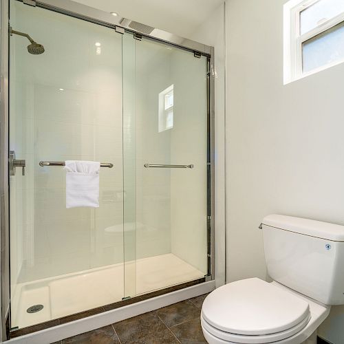 A bathroom with a glass-enclosed shower, a wall-mounted towel rack inside, and a white toilet beneath a small window.