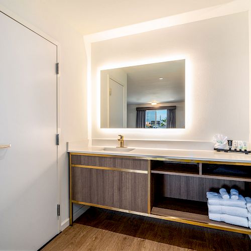 A modern bathroom features a backlit mirror, a sink with a gold faucet, a wooden vanity with towels and toiletries, and a door on the left.