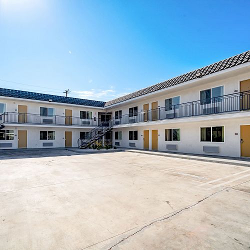 An exterior view of a two-story motel with a large empty courtyard, featuring multiple doors and windows, under a clear blue sky.