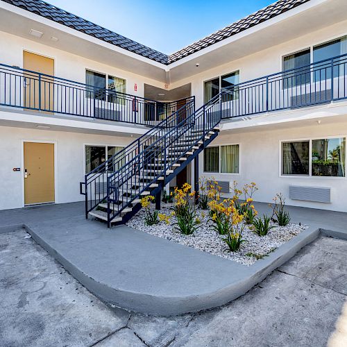 An exterior view of a two-story motel with a central staircase, yellow doors, and a small garden area with plants.