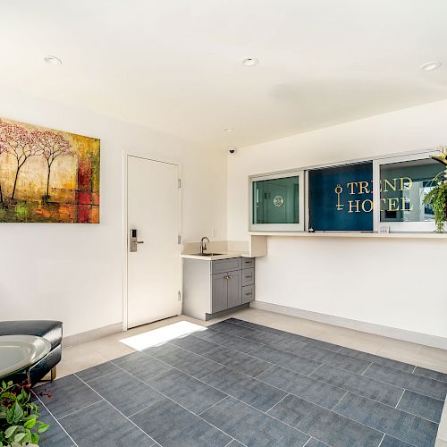 A well-lit reception area featuring a modern chair, a coffee table, plants, a wall painting, and a service window with the text 