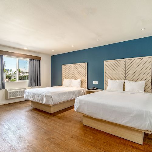 A hotel room with two double beds, wooden floors, blue accent wall, window with drapes, and a chair near the air conditioning unit.