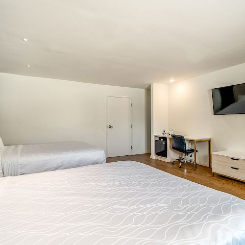 The image shows a clean, modern hotel room with two beds, a TV, a desk with a chair, and a small fridge below the desk.