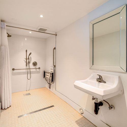 A spacious, accessible bathroom with a walk-in shower, handrails, a sink, and a mirror. The shower area has a curtain and bench.