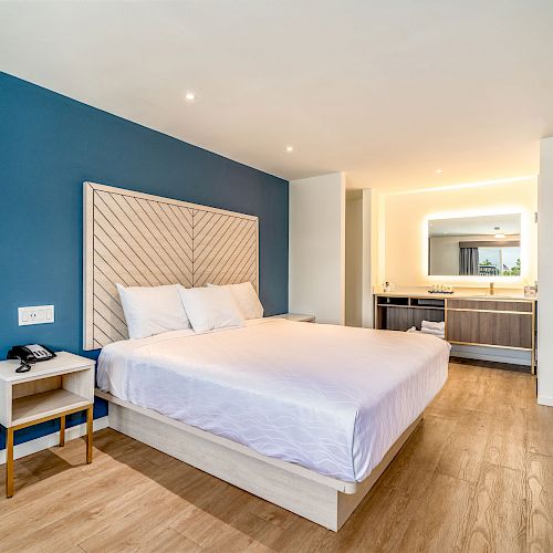 A modern hotel room with a blue accent wall, a large bed, nightstand, and a minimalist decor style. A well-lit bathroom is visible in the background.