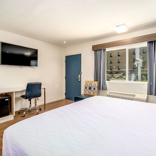 A clean, modern hotel room features a bed, wall-mounted TV, desk with chair, mini fridge, blue curtains, and a window displaying an outside building view.