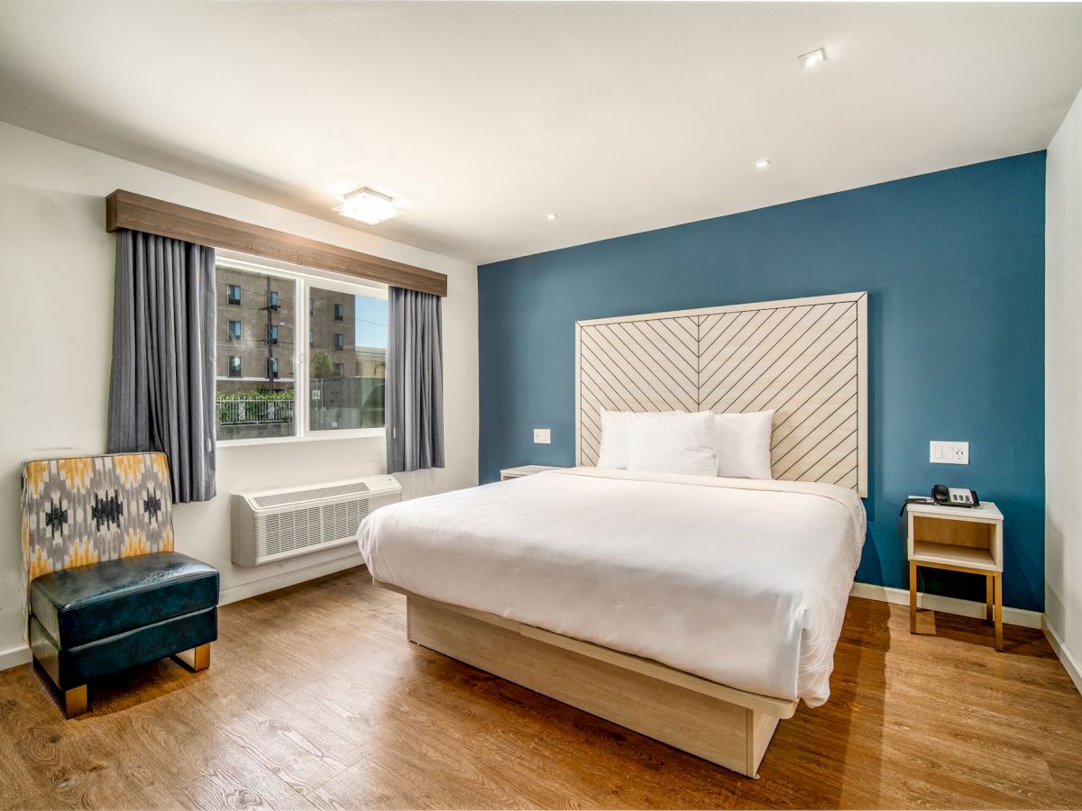 A modern hotel room featuring a large bed, wooden floor, blue accent wall, window with curtains, chair, and nightstands.