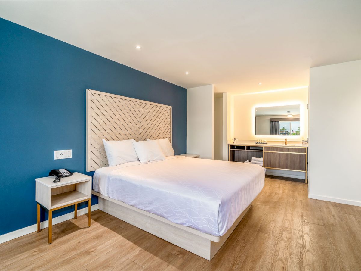 A spacious bedroom with a light-colored bed, blue accent wall, side table, and an ensuite bathroom featuring a lit mirror and modern fixtures, ending the sentence.