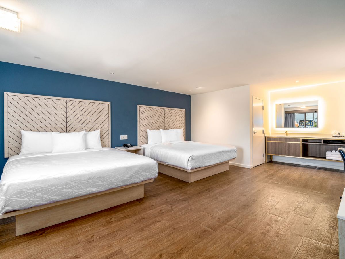 A modern hotel room with two large beds, wooden floor, blue accent wall, bathroom with large mirror, and a desk area.