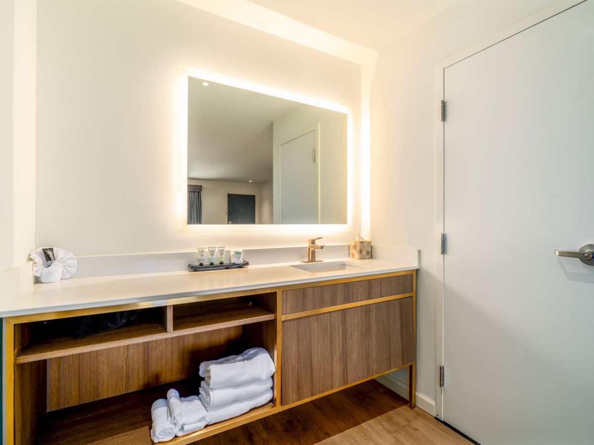 A modern bathroom vanity area with a backlit mirror, sink, toiletries, towels on open shelves, and a closed white door to the right.