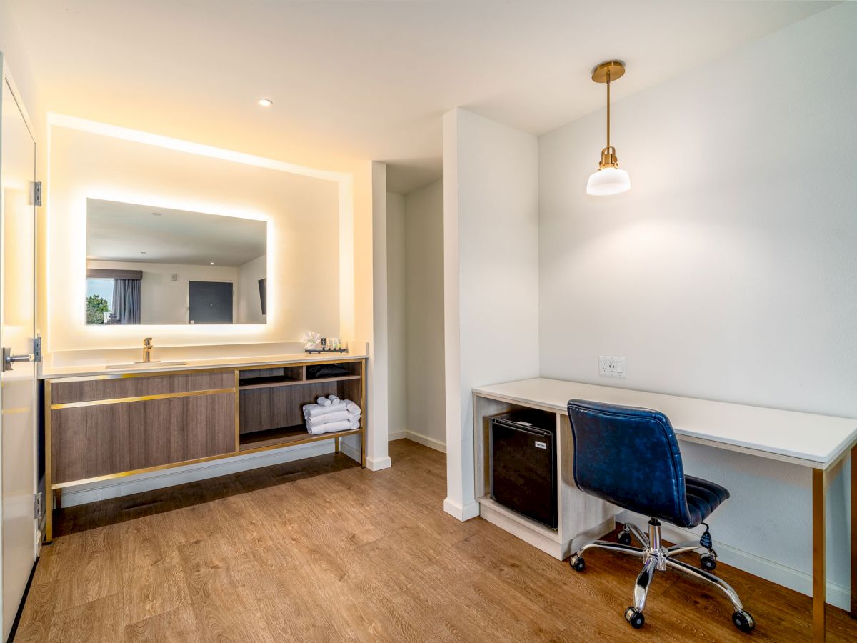 A modern room with a wooden floor, vanity with illuminated mirror, desk with blue chair, and hanging light fixture.