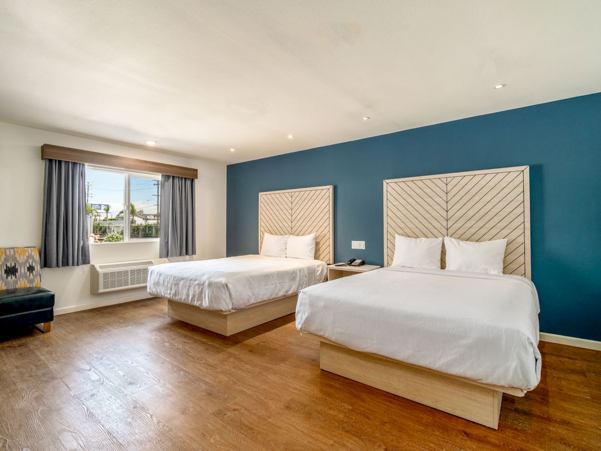 A hotel room with two double beds, a window with curtains, a patterned chair, and wooden flooring. The wall behind the beds is painted blue.