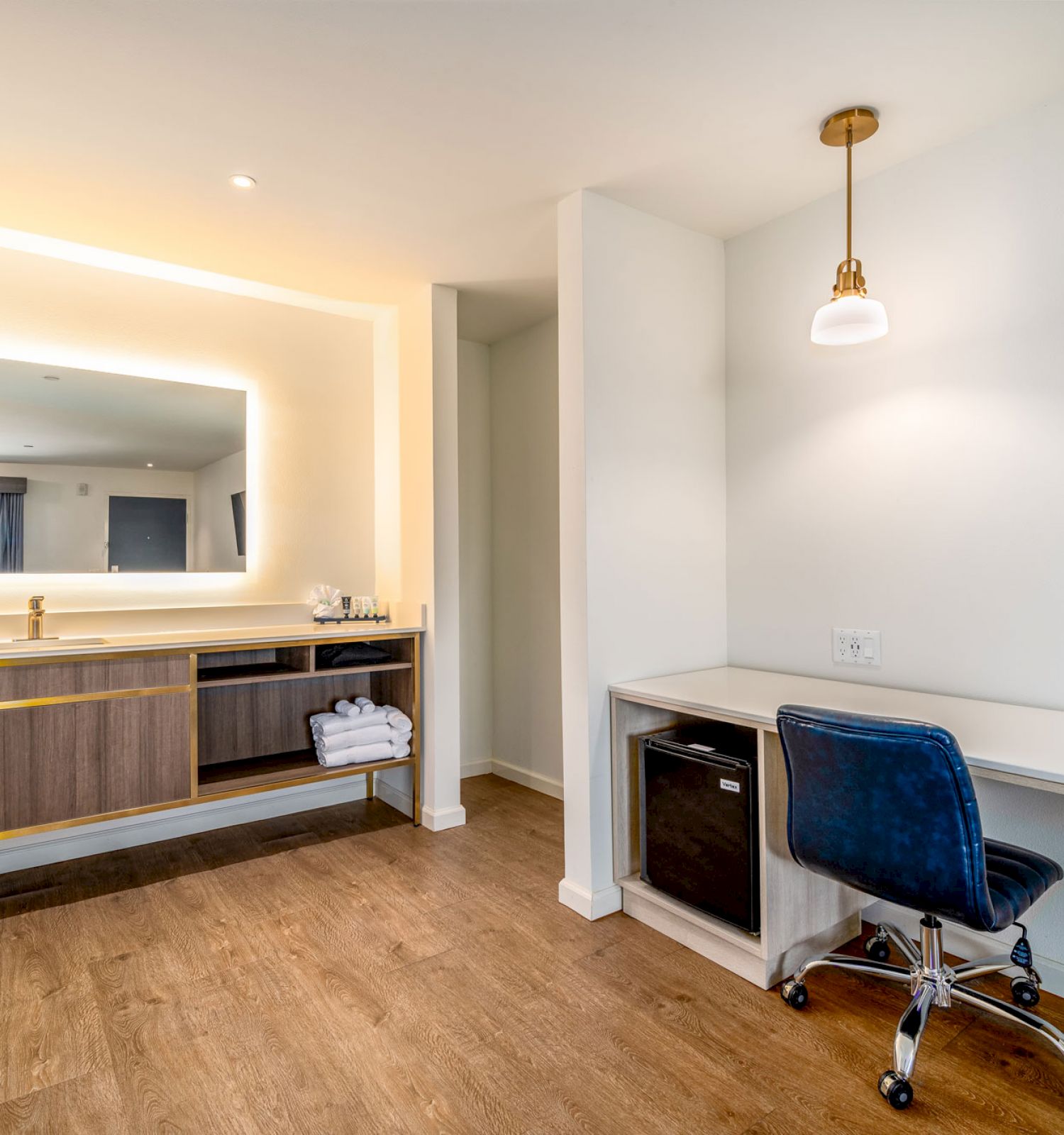 A modern room with a wooden floor, a vanity with a lighted mirror, a desk with a blue chair, and a pendant light above the desk.