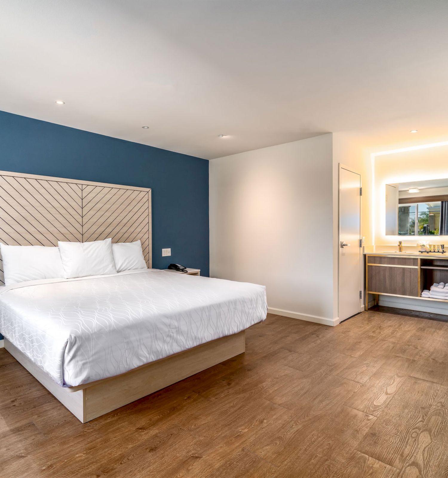 The image shows a modern hotel room with a large bed, nightstands, a desk with a chair, and an open bathroom setup.