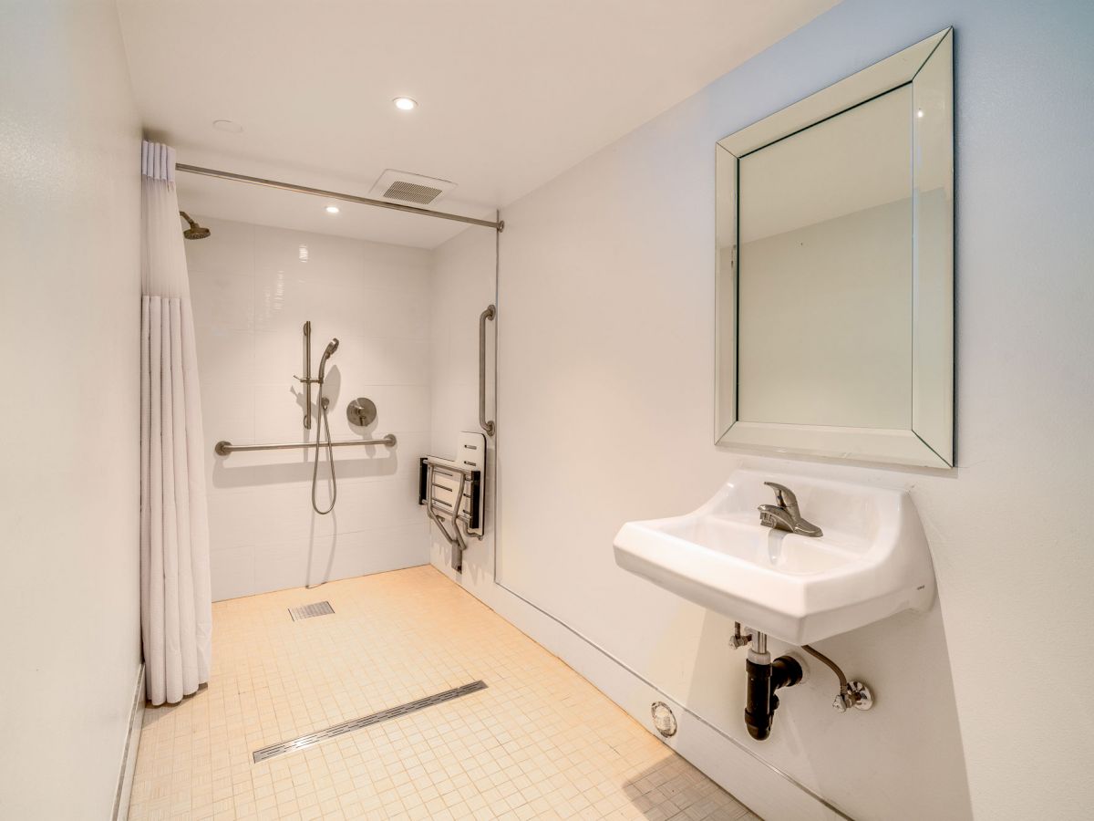 A bathroom with a wall-mounted sink, mirror, and a spacious shower area with grab bars and a shower seat, designed for accessibility.