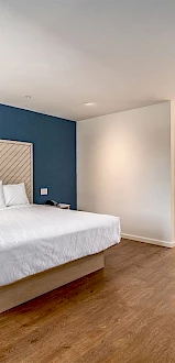 A modern hotel room with a large bed, blue accent wall, nightstand, a desk with a chair, and an open washroom with a lit mirror above the sink.