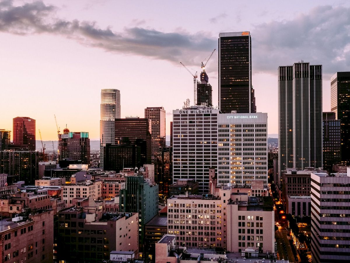 This image shows a city skyline at dusk, featuring tall buildings, a few cranes, and cloudy skies with a hint of sunset colors in the background.