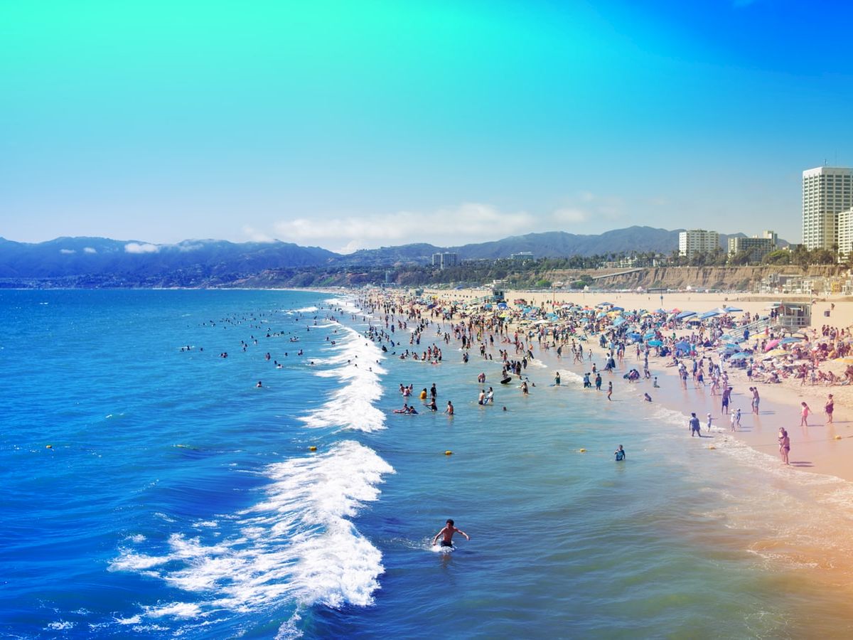 A crowded beach scene with people swimming and relaxing on the sand, buildings and hills visible in the background, under a clear blue sky.