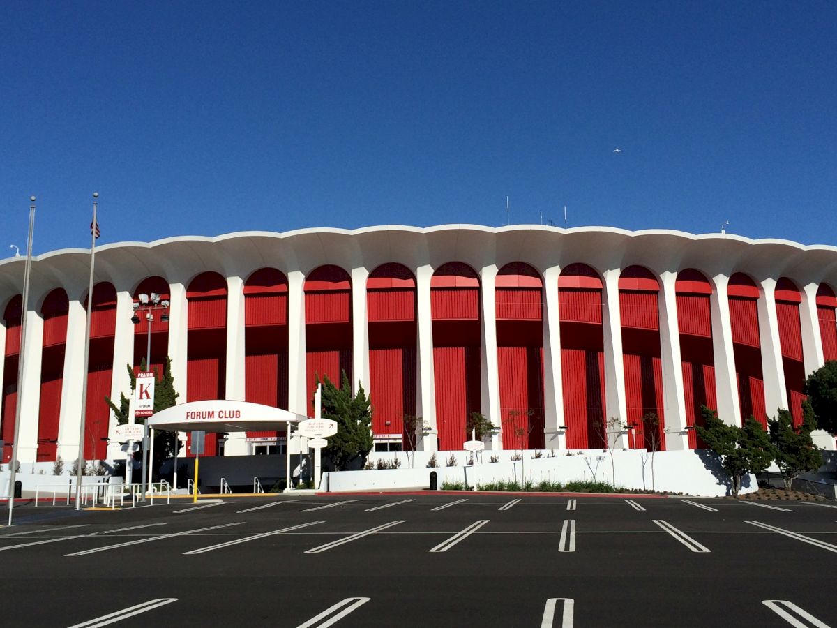 The image shows a large, round building with a red and white color scheme, numerous columns, and a sign that reads “Forum Club,” with a blue sky above.