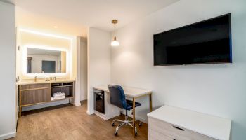 The image shows a modern hotel room with a large wall-mounted TV, a desk with a chair, a dresser, and a bathroom vanity with a mirror and towels.