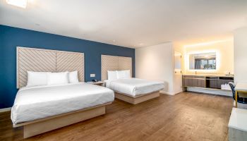 The image features a modern hotel room with two large beds, a wooden floor, a blue accent wall, and a vanity area with a mirror and lights.