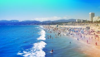 People are enjoying a sunny day at a crowded beach with waves crashing. Buildings and mountains are visible in the background, under a blue sky.