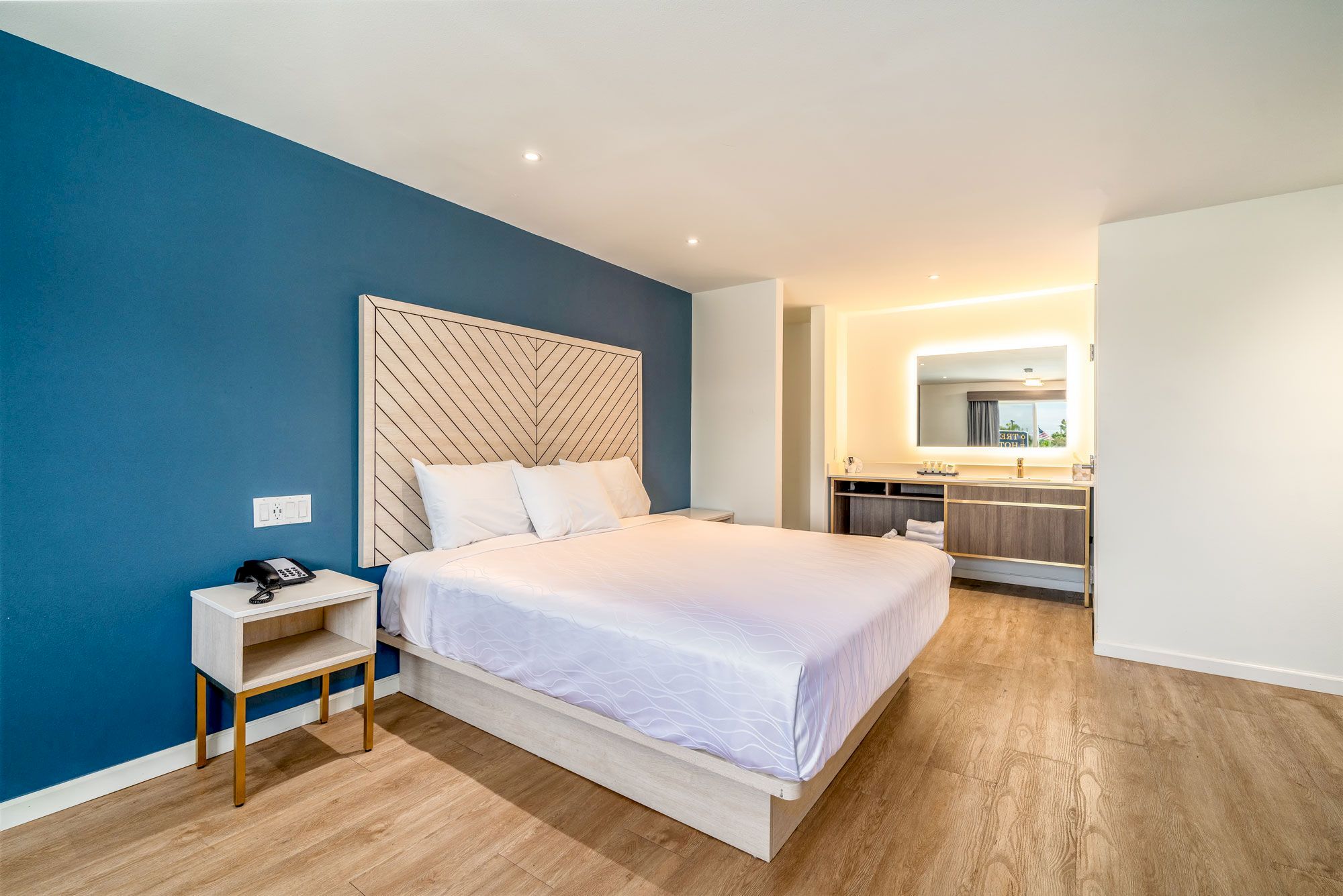 A modern bedroom with a large bed, blue accent wall, wooden floor, bedside table, and an open en-suite bathroom with illuminated mirror.