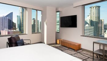 The image shows a modern hotel room with large windows offering a city view, a bed, a chair, a wall-mounted TV, and minimalist furniture.