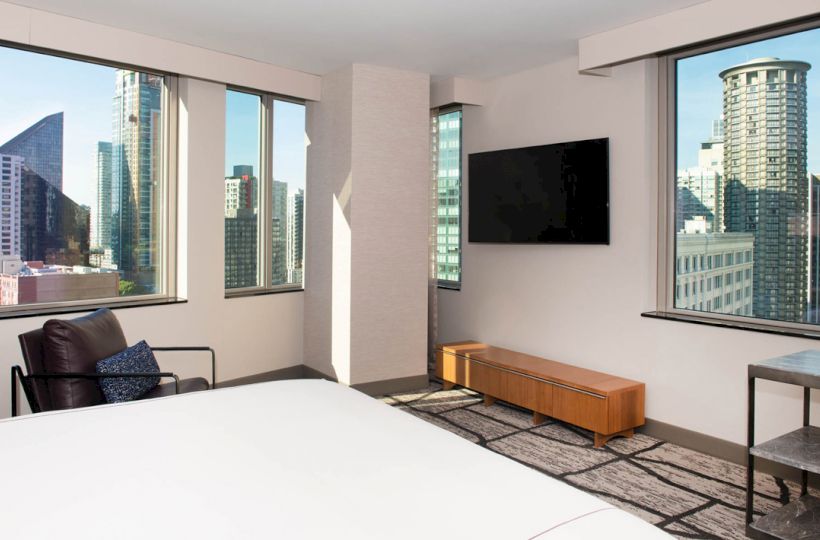 The image shows a modern hotel room with large windows offering a city view, a bed, a chair, a wall-mounted TV, and minimalist furniture.