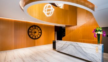 Modern reception area with a marble desk, wooden accents, large wall clock, and a stylish pendant light fixture hanging from a spiral ceiling.