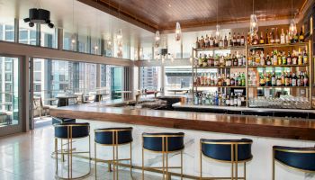 This image shows a modern bar with a wooden counter, blue and gold bar stools, and shelves stocked with various bottles of liquor. The sentence ends.