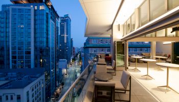 A modern cityscape at dusk with towering buildings and a well-lit, furnished balcony overlooking a street from a high floor.