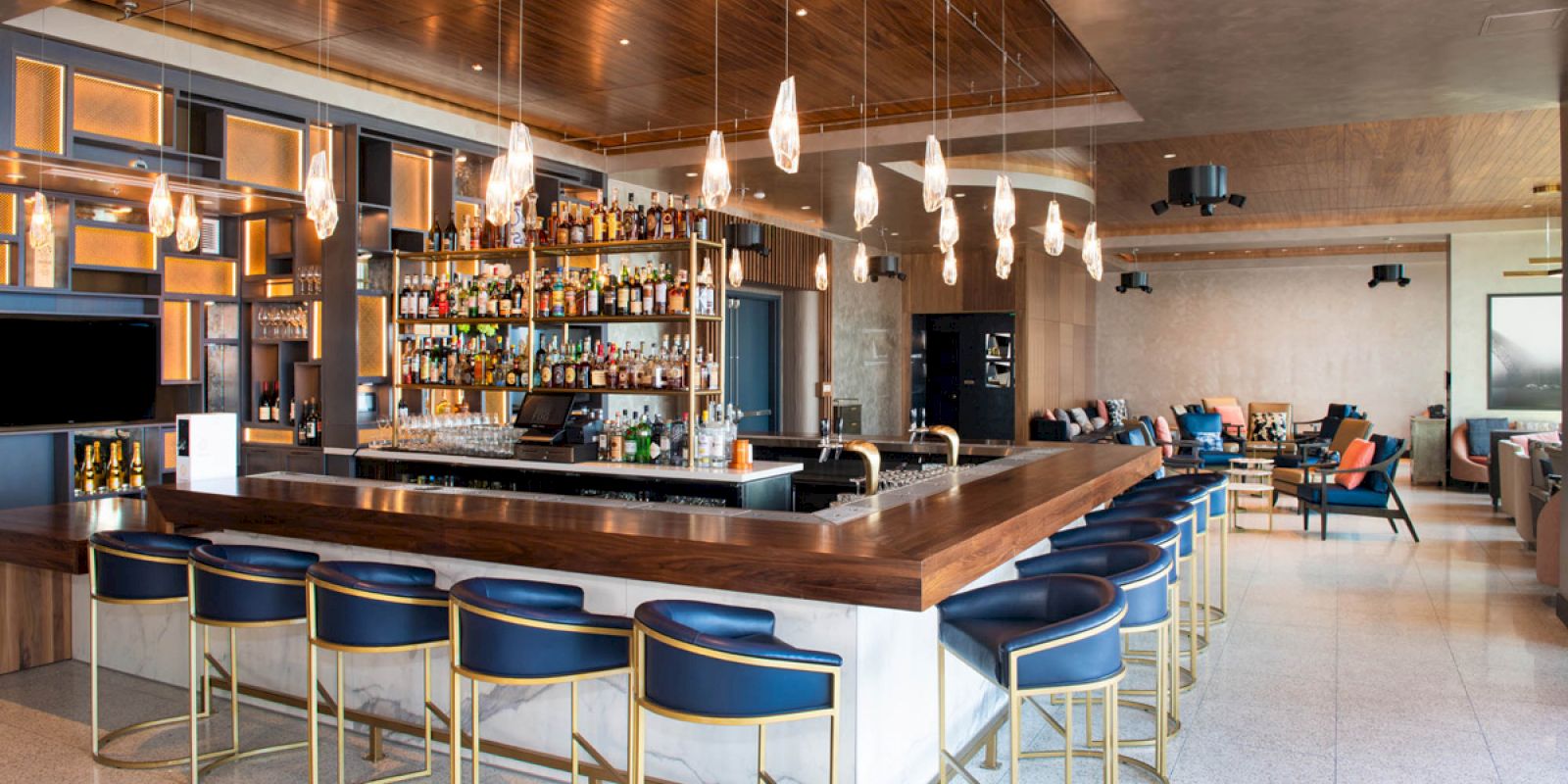 A modern bar area with a well-stocked liquor display, stylish seating, pendant lighting, and a lounge area in the background.