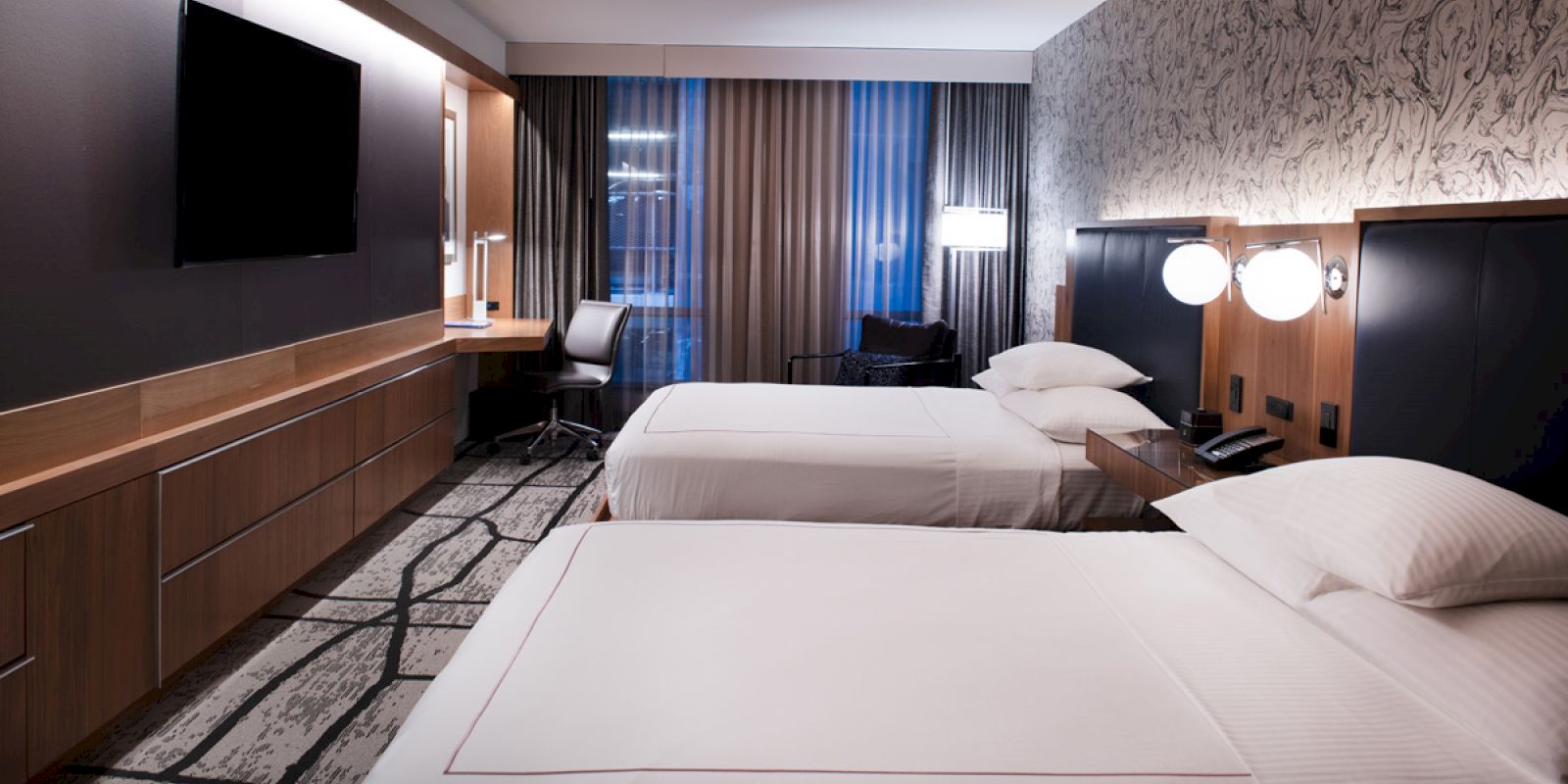 A modern hotel room with two neatly made beds, a flat-screen TV, a desk with a chair, and large windows covered by curtains.