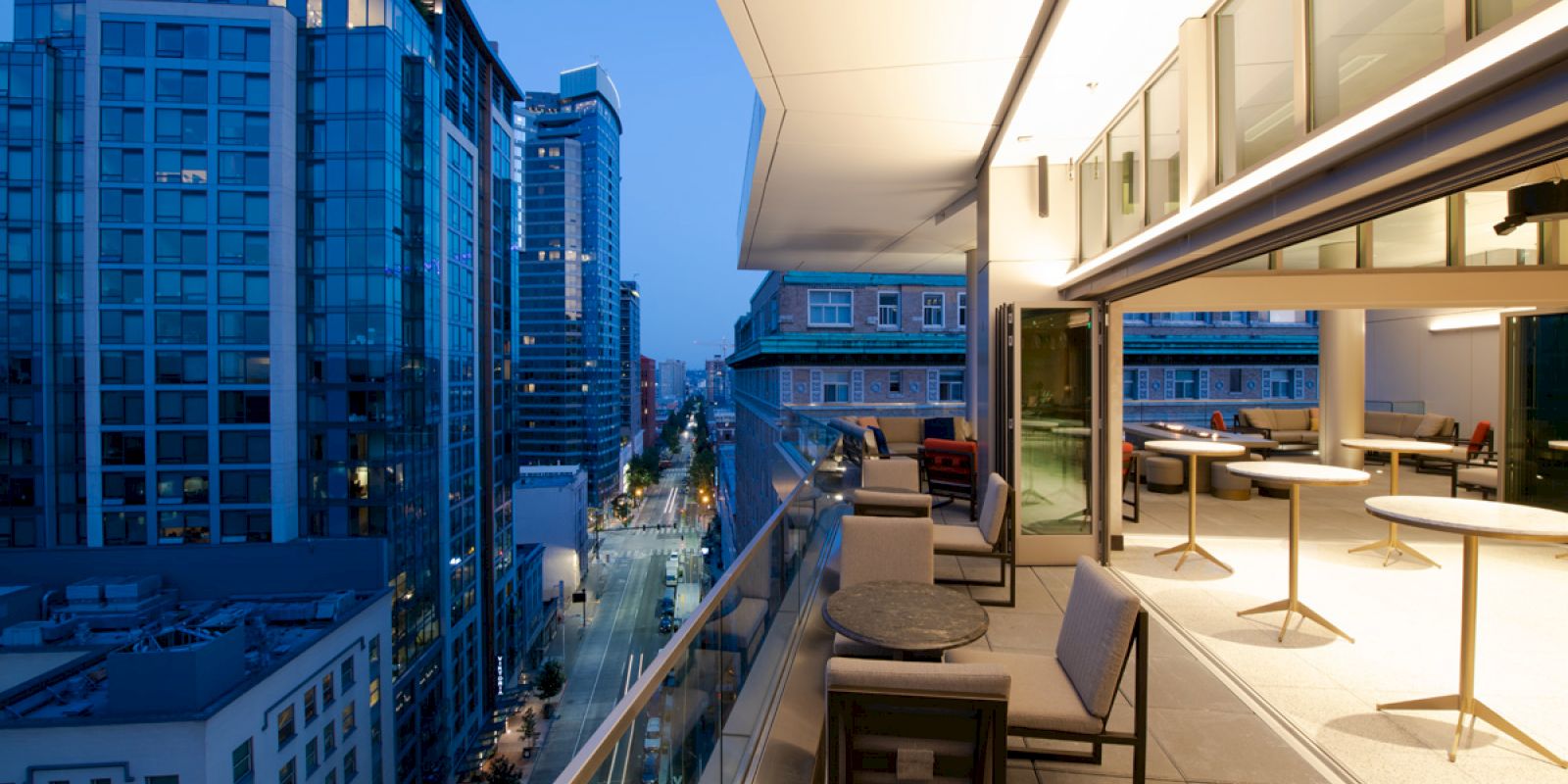 This image shows a modern balcony with seating and tables, overlooking a city street lined with tall buildings at dusk.