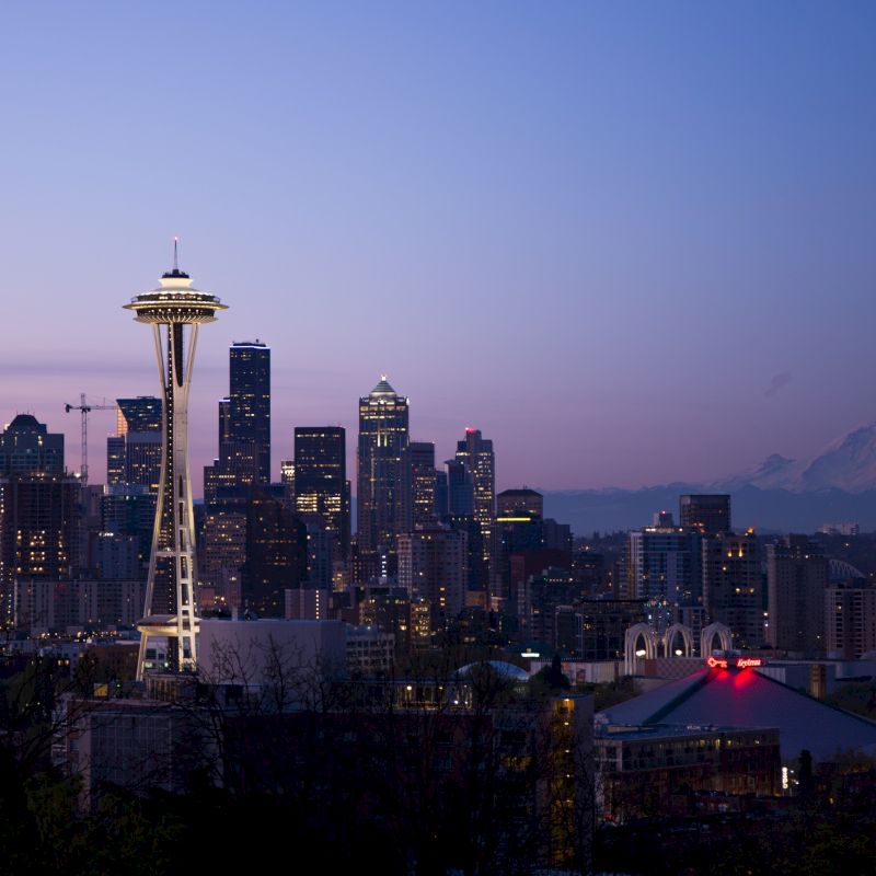 The image shows the Seattle skyline at dusk, featuring the Space Needle prominently, with Mount Rainier faintly visible in the background.