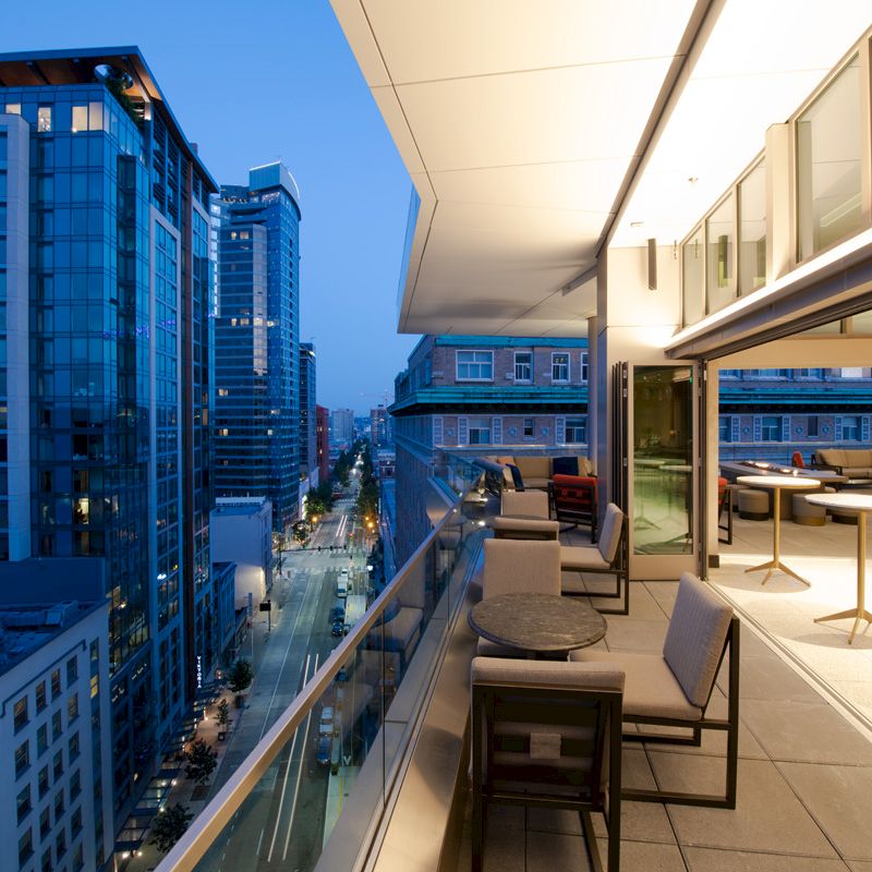 This image shows a modern cityscape with tall buildings, viewed from a spacious outdoor balcony featuring tables and chairs, during twilight.