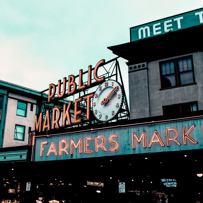 The image shows an iconic sign for a public market and farmers market with a clock, set against urban buildings.