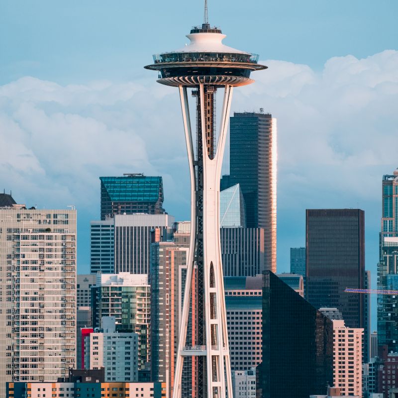 The image shows the Space Needle with a backdrop of various buildings and skyscrapers under a partly cloudy sky, highlighting an urban landscape.