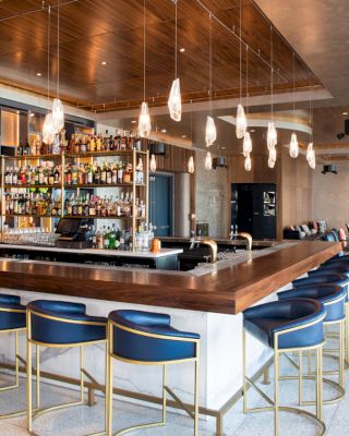 This image shows a modern bar with a wooden counter, high chairs, hanging lights, and a variety of bottles. The setting is stylish and comfortable.